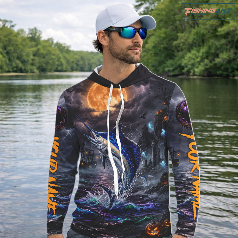 A man wearing a black baseball cap and sunglasses stands on a boat holding a fishing rod, wearing a custom all-over print hoodie featuring a blue marlin, a full moon, and orange text on the sleeves.