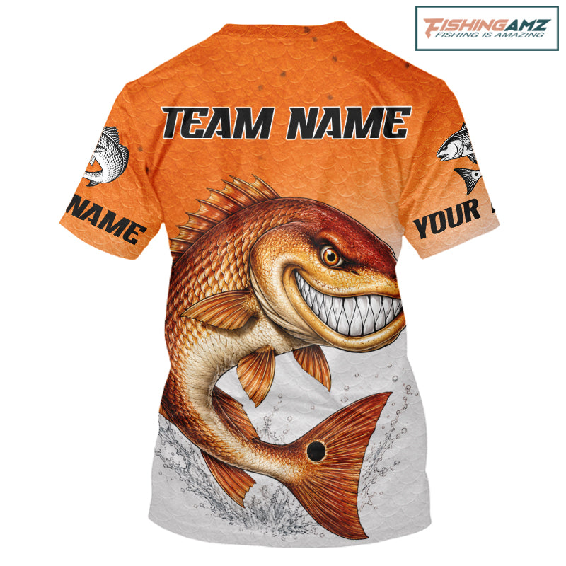 An orange short-sleeve t-shirt with a large graphic of a smiling bass fish on the front, customizable text placeholders 'TEAM NAME' and 'YOUR NAME' on the chest and sleeves, and a 'FishingAMZ' logo in the top right corner.