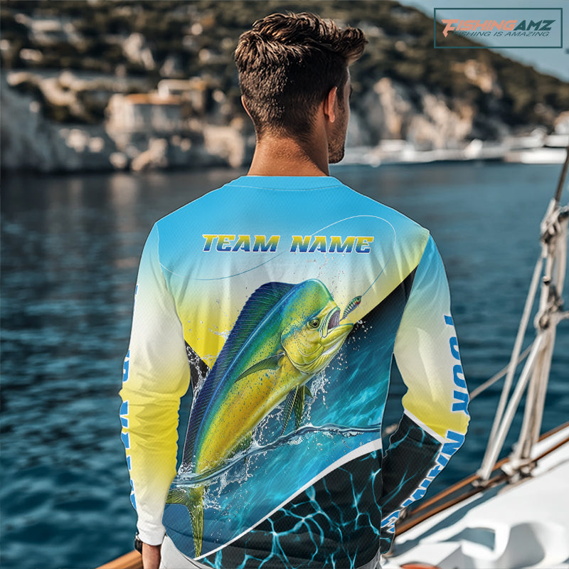 A man viewed from behind wearing a long-sleeve graphic fishing shirt with a large mahi-mahi fish print and 'TEAM NAME' text on the back, standing at a beach with the ocean in the background.