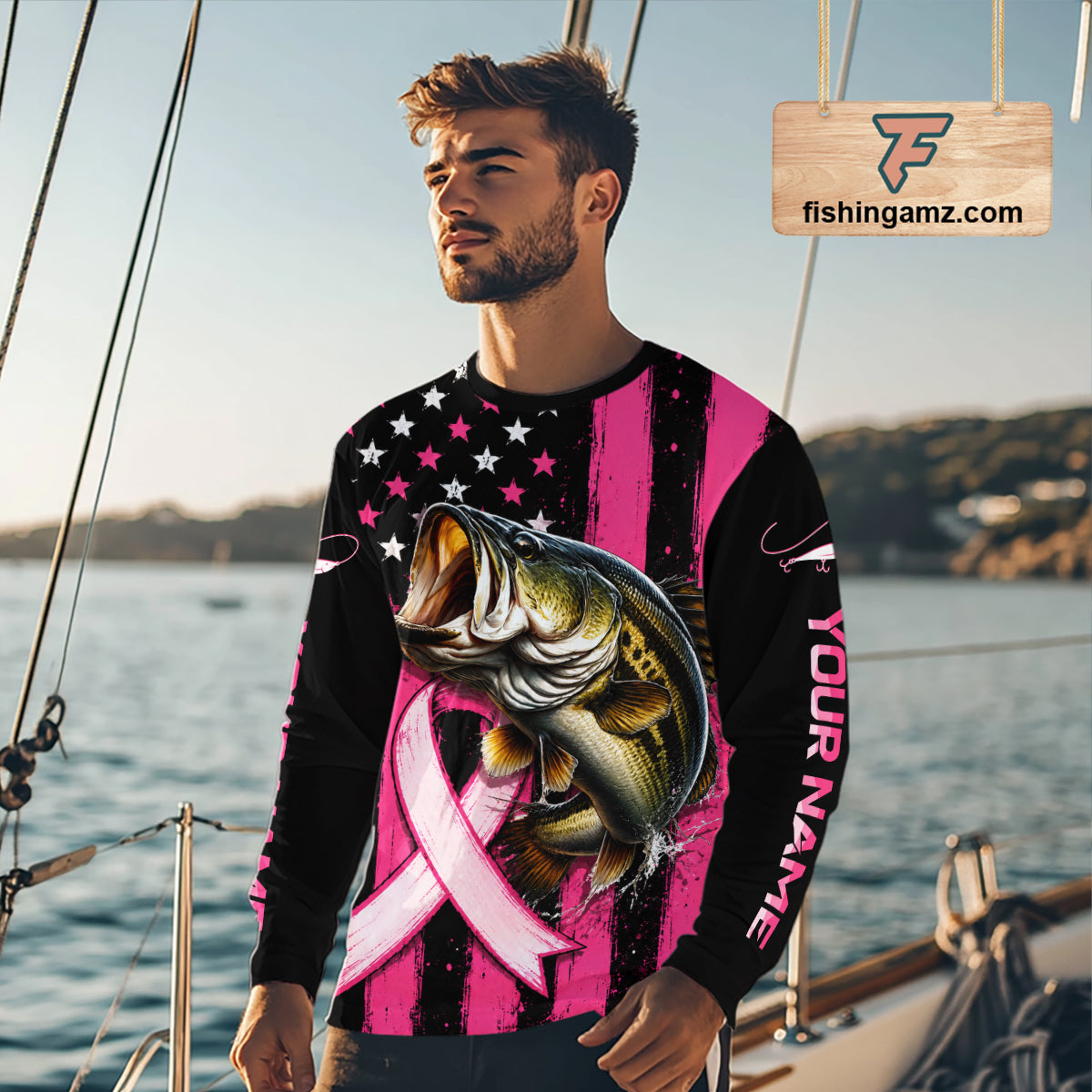 A man wearing a long-sleeve custom shirt with a pink and black striped design, a black American flag pattern on the upper chest, a large bass graphic over a pink ribbon, and 'YOUR NAME' printed down one sleeve. The background shows a blurred marina setting with boats and water.