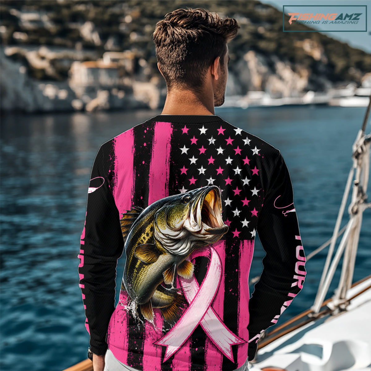A man wearing a long-sleeve custom shirt with a pink and black striped design, a black American flag pattern on the upper chest, a large bass graphic over a pink ribbon, and 'YOUR NAME' printed down one sleeve. The background shows a blurred marina setting with boats and water.