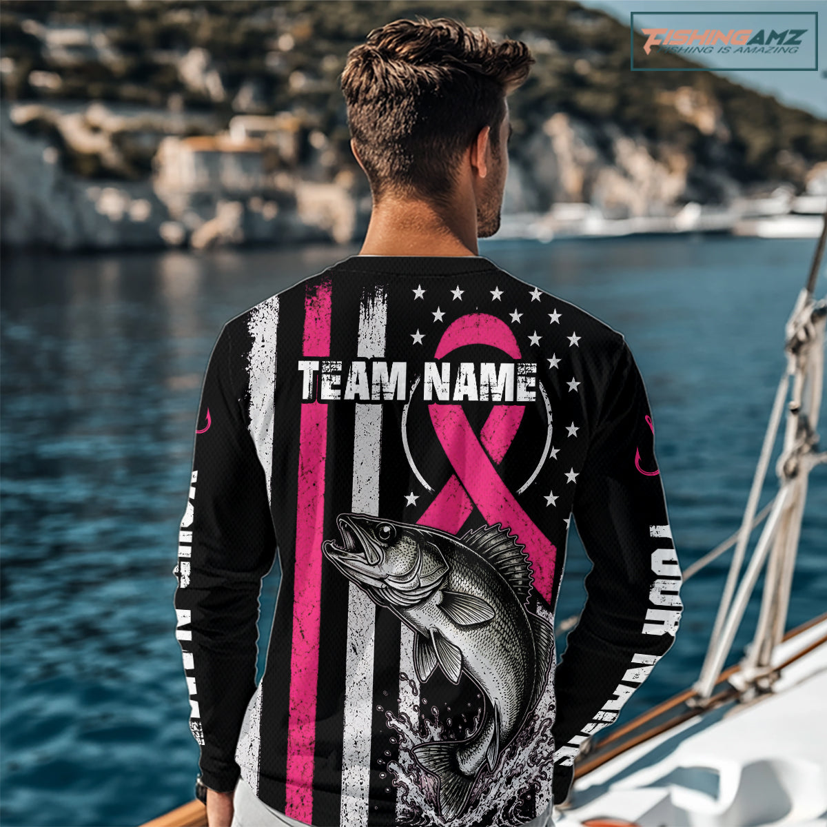 A man standing on a beach viewed from behind, wearing a black long-sleeve shirt with a large pink and white graphic design featuring a fish, stars, stripes, and customizable text. The background shows the blue ocean and sky.