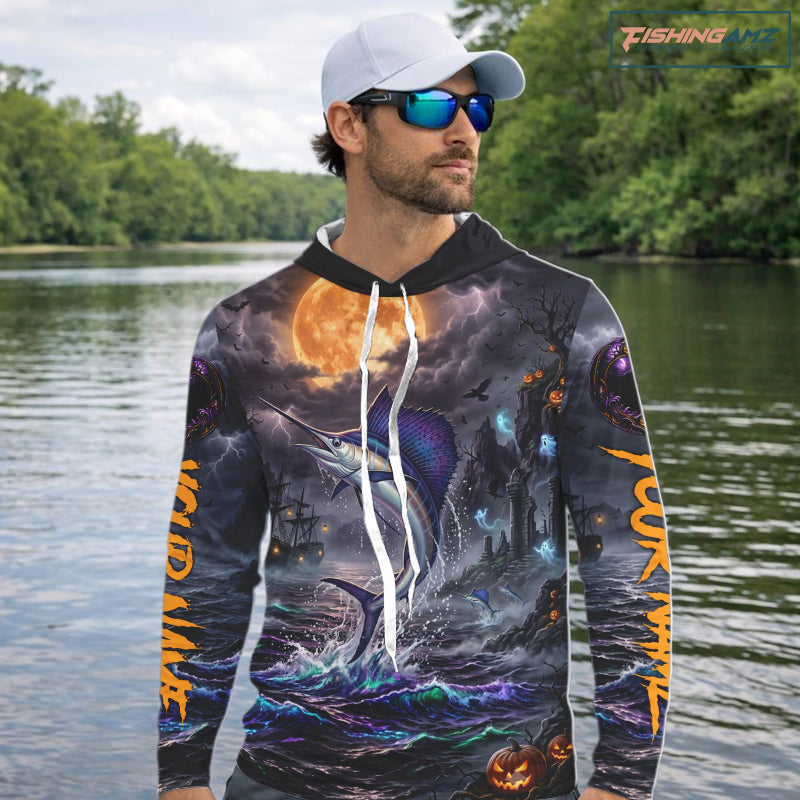 A man wearing a black baseball cap and sunglasses stands on a boat holding a fishing rod, wearing a long-sleeve hoodie with an all-over Halloween-themed graphic print featuring a fish, a ghost ship, jack-o'-lanterns, and orange text on the sleeves.