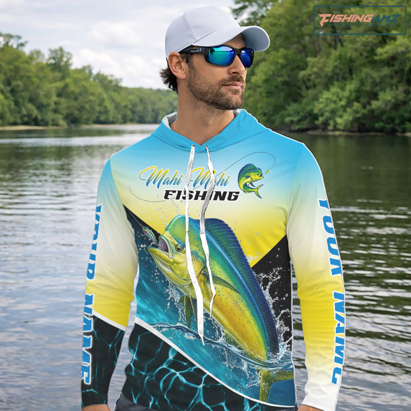A man wearing a custom blue and yellow gradient fishing hoodie with a large mahi-mahi fish graphic, sunglasses, and a black cap, standing on a boat with fishing rods and the ocean in the background. 