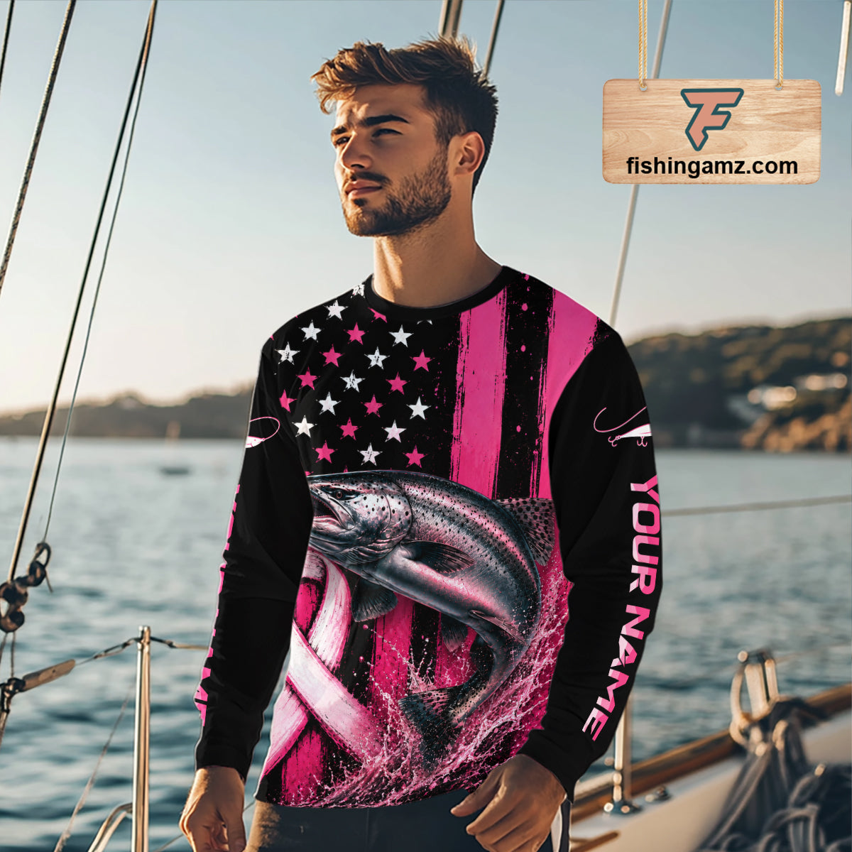 A man wearing a long-sleeve custom crewneck shirt with a pink and black American flag design, a large fish graphic, and 'YOUR NAME' printed on the sleeve, standing on a boat with water and another boat in the background.