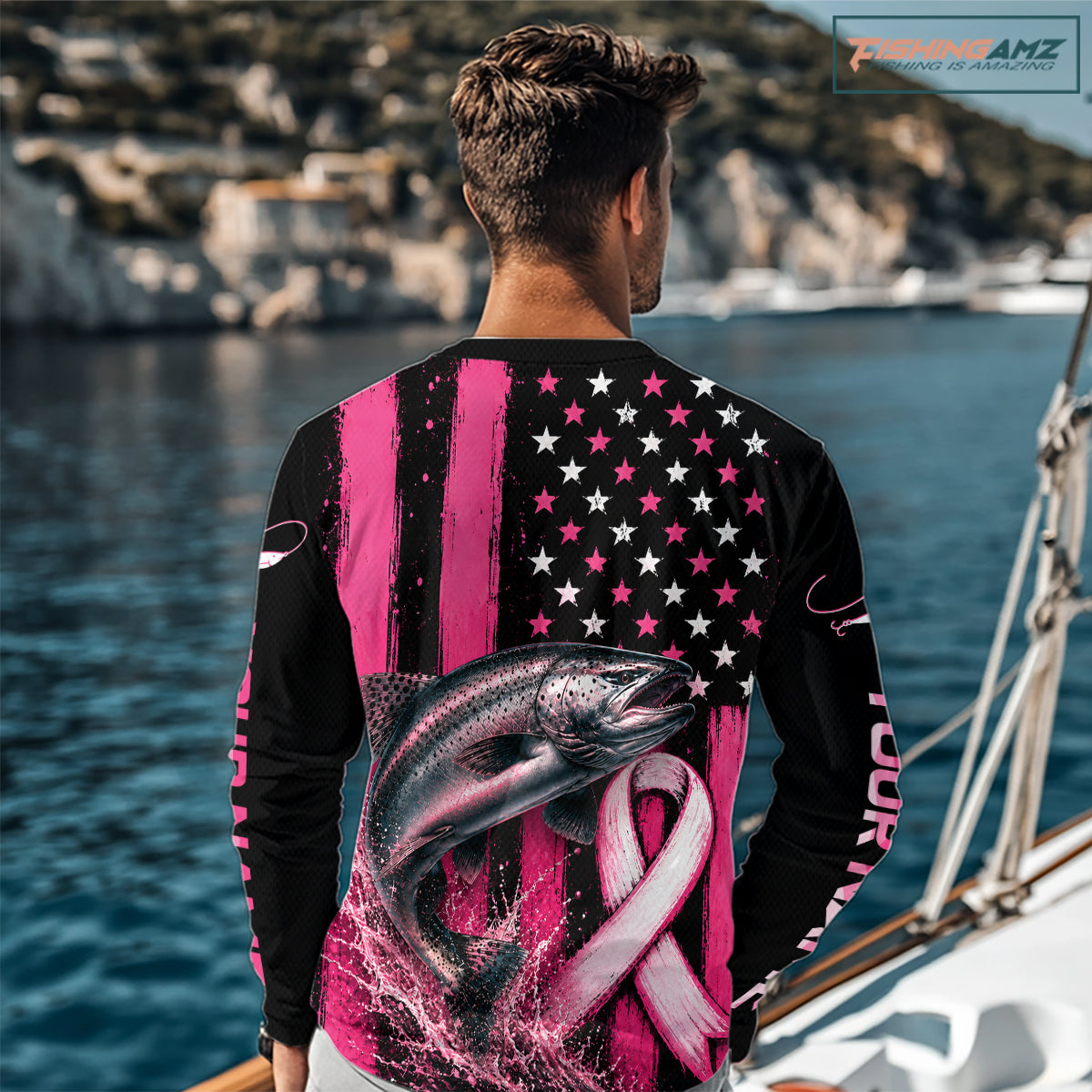 A man viewed from behind wearing a long-sleeve black shirt with a pink and white patriotic fish graphic, stars-and-stripes pattern, and pink ribbon design, standing by the ocean. The shirt includes the text 'YOUR NAME' on the sleeve.