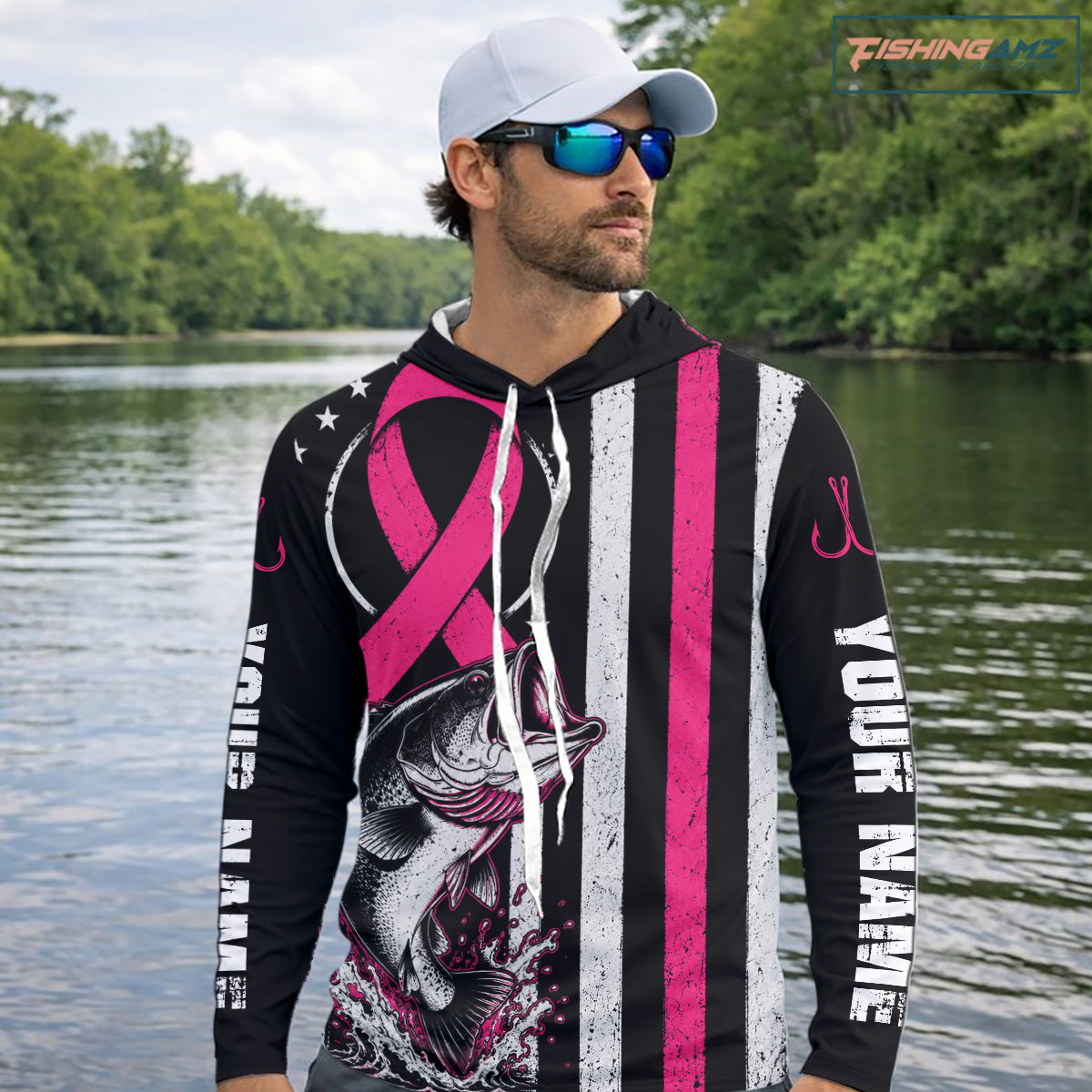 A man on a boat wearing a black custom hoodie with pink and white stripes, a fish graphic, and white text reading 'YOUR NAME'. He is also wearing a dark cap and sunglasses, with fishing rods and water in the background.