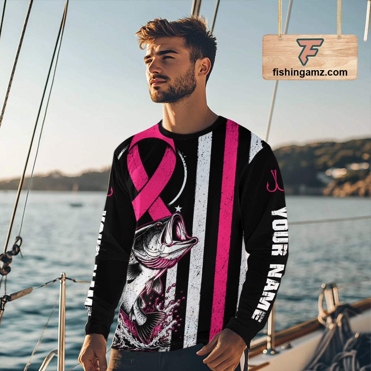 A man wearing a long-sleeve black and white distressed graphic shirt with a pink ribbon, a fish illustration, and 'YOUR NAME' printed on the sleeve, standing on a boat with water and another boat in the background.