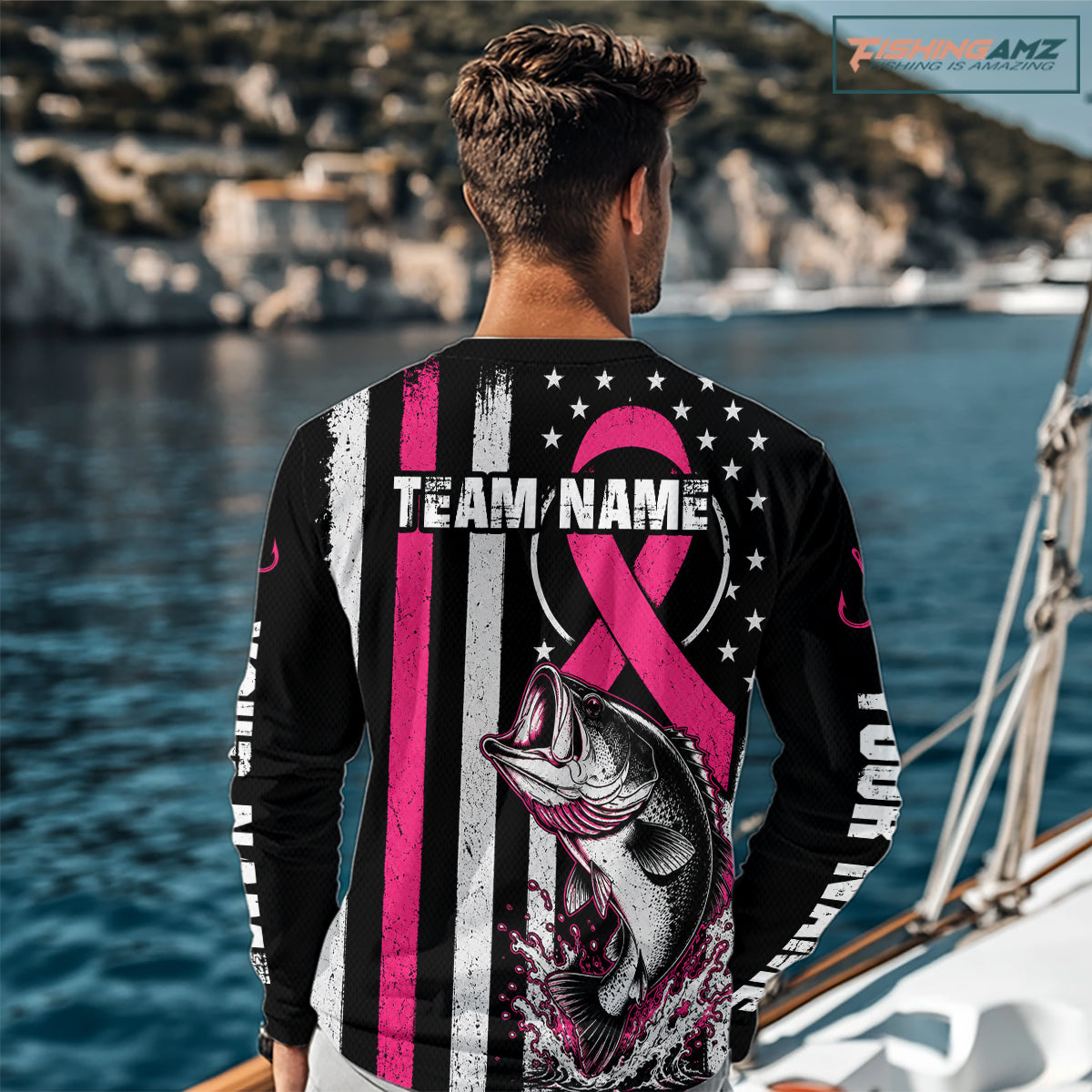 A man stands on a beach facing the ocean, wearing a black long-sleeve graphic shirt with a pink ribbon, fish illustration, and customizable text. A 'FISHINGAMZ watermark is visible in the top right corner.