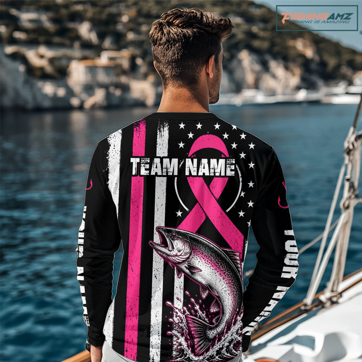 A man stands on a beach facing away from the camera, wearing a black long-sleeve shirt with a large pink and white graphic design featuring a fish, stars, stripes, and customizable text. The background shows the blue ocean and sky.